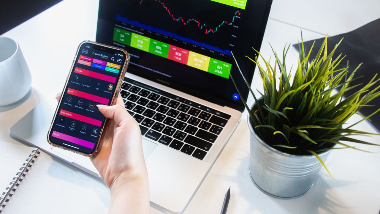 A person using a smartphone app to trade stocks with a laptop displaying market data.